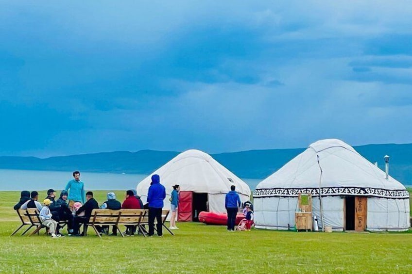 A group of people gathered around yurts in a grassy field with mountains in the background. 1