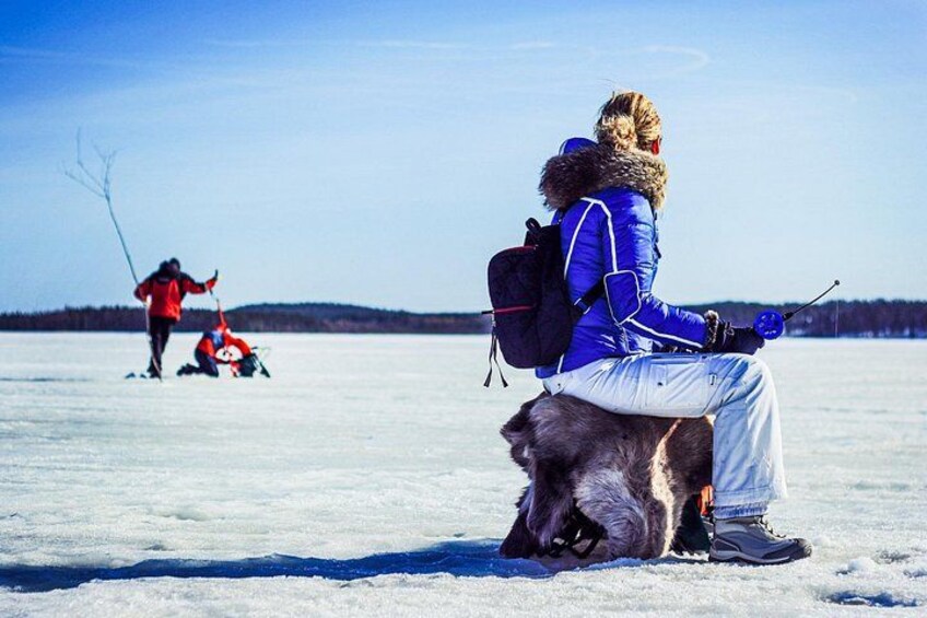 A person sitting on a sled with a dog, another person fishing in the background, and a frozen landscape. 3