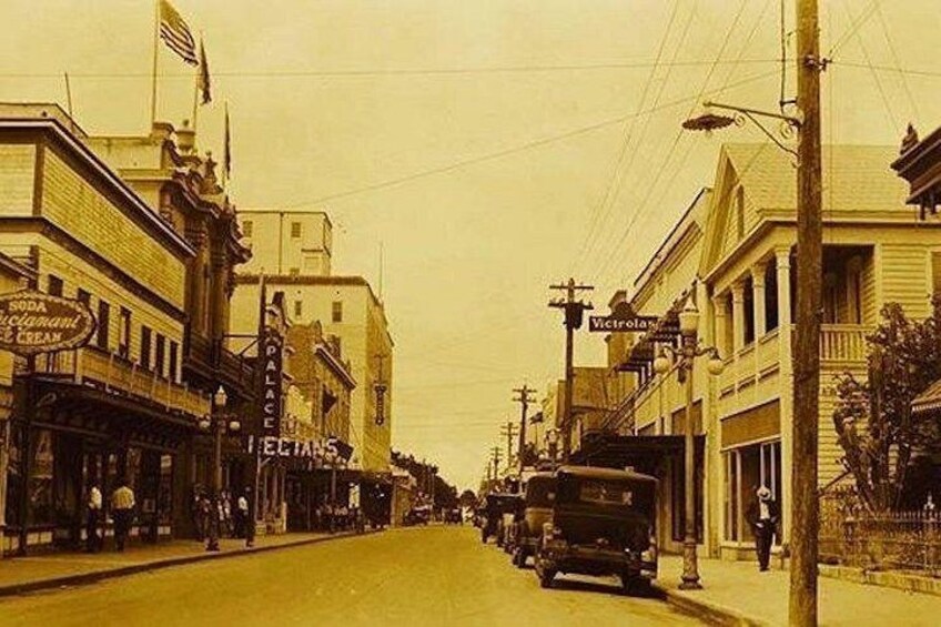 Une scène de rue d’époque, avec des bâtiments, une voiture ancienne et une enseigne indiquant « Soda Fountain & Creamery ». 1