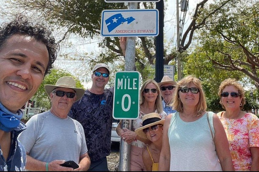 A group of people standing near a signpost with a 'MILE 0' marker and an 'AMERICAS BYWAYS' sign. 2