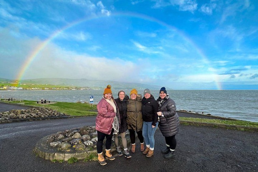 Un groupe de personnes se tient sur un sentier rocheux, au bord d’une étendue d’eau, avec un arc-en-ciel en arrière-plan. 2