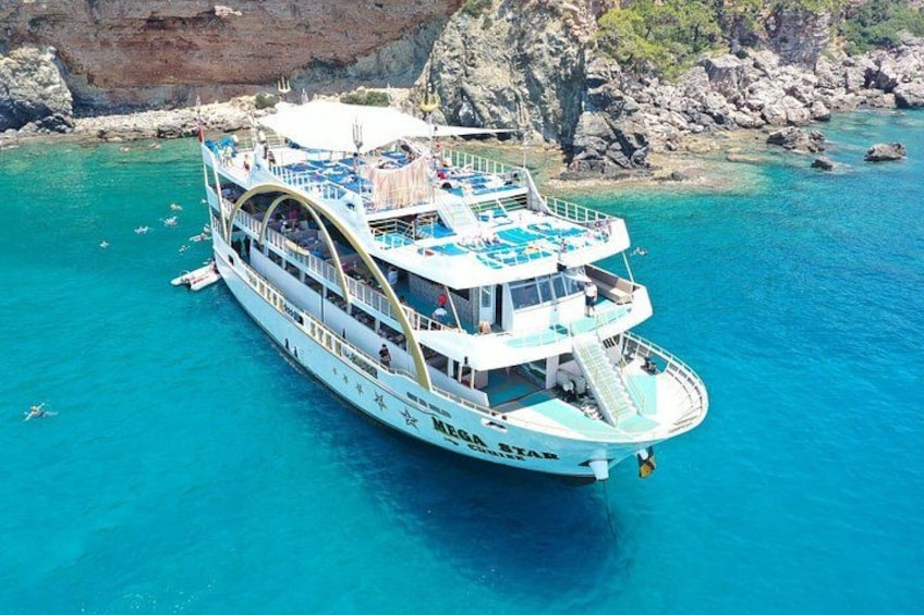 Een grote witte boot met meerdere dekken en een blauw zonnescherm, drijvend in helder turquoise water nabij rotsachtige kliffen. 2