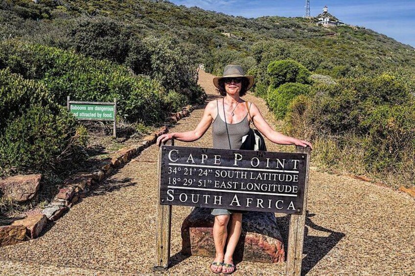 Eine Person steht hinter einem Schild am Kap der Guten Hoffnung in Südafrika, auf dem Koordinaten und eine Warnung bezüglich Paviane abgebildet sind. 3