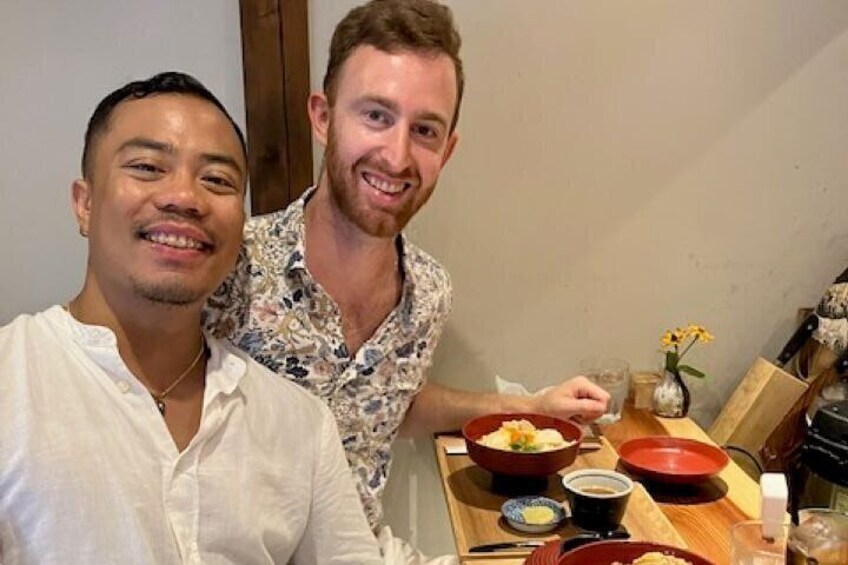 Two people sitting at a table with bowls of food and a vase with flowers. 1