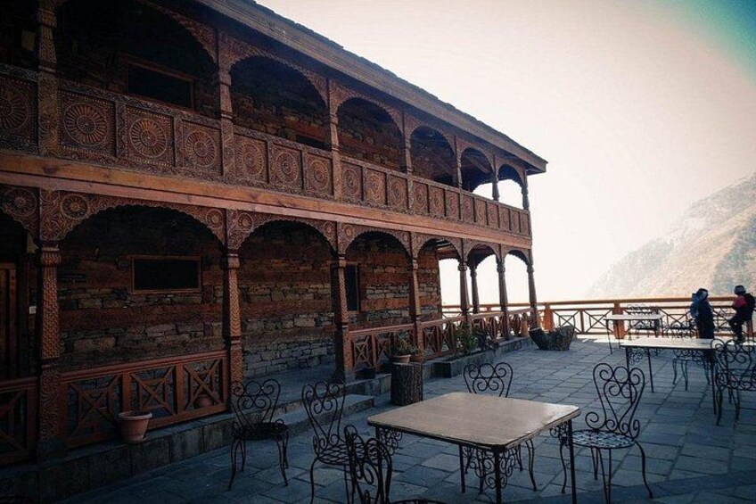 Een klassiek gebouw met gedetailleerd baksteenwerk en bogen, een terras met tafels en stoelen, en een bergachtige achtergrond. 5