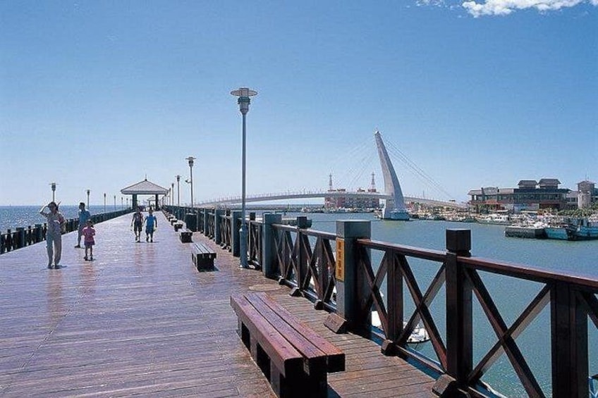 A wooden pier extending into the water with benches and a gazebo. A bridge and buildings are visible in the background. 1