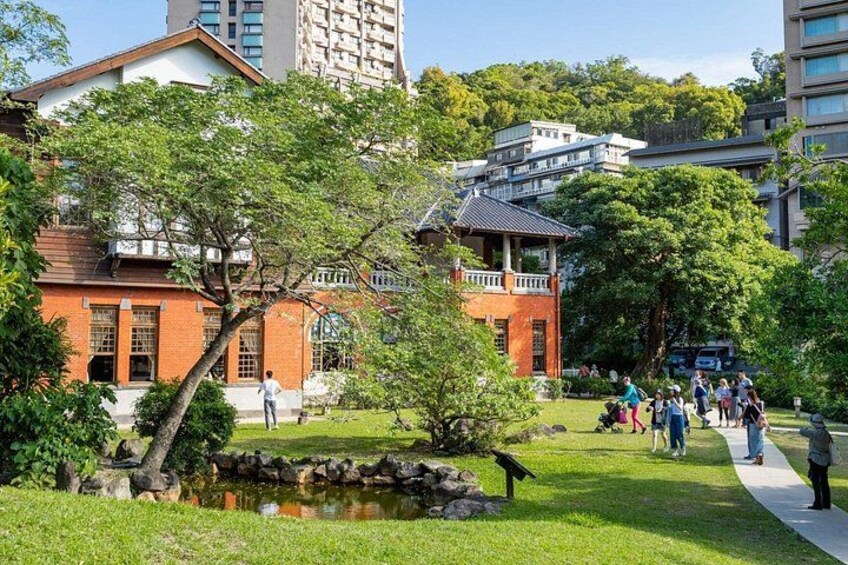 A park with a pond, trees, a red building, and people walking. 1