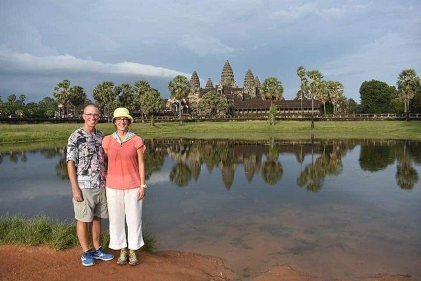 Two people standing by a calm water body with a reflection, in front of a large temple complex with multiple towers. 2
