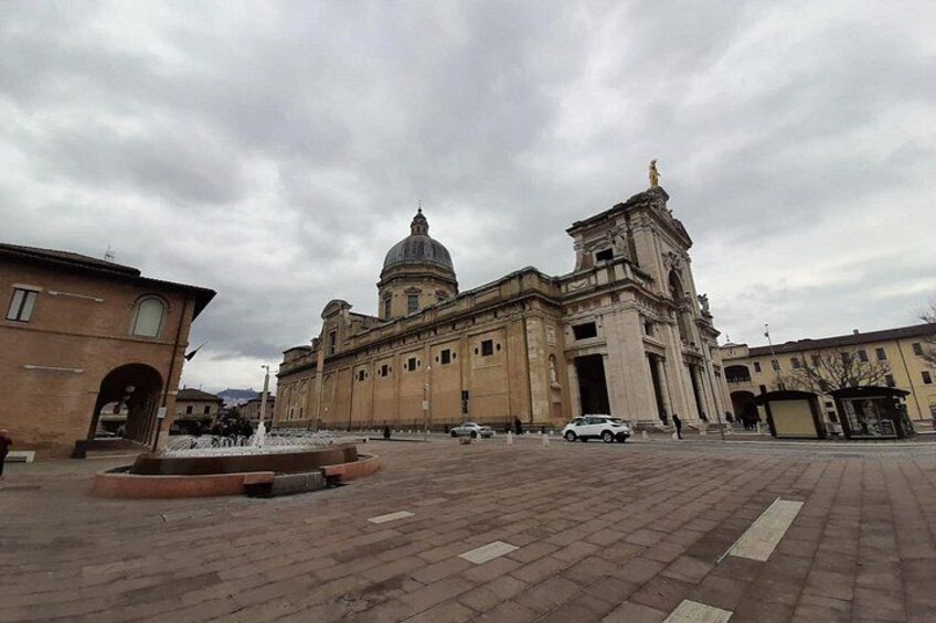 A large historic building with a dome and two towers, a paved square with a fountain, and a few parked cars. 2