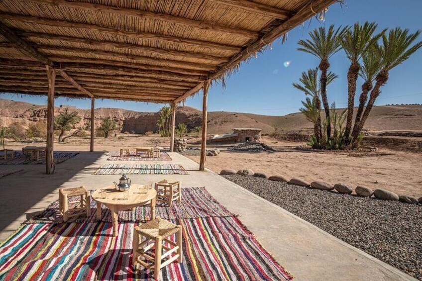 A covered outdoor dining area with colorful rugs, wooden furniture, and palm trees in a desert landscape. 3