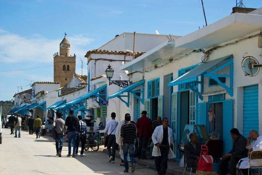 Una calle concurrida con gente caminando, tiendas con toldos azules y una torre destacada al fondo. 2