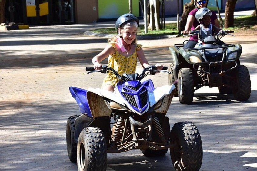 A child riding a blue ATV on a paved road with trees and a building in the background. 5