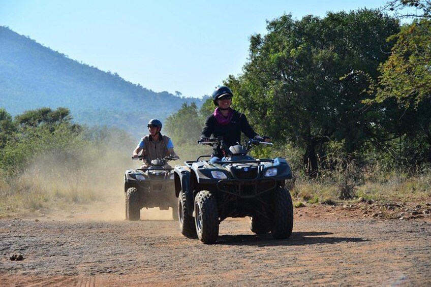Two people riding ATVs on a dirt road with trees and hills in the background. 3
