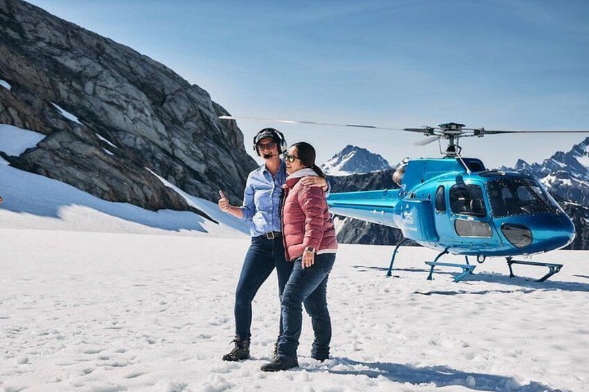 Two people standing on snow-covered ground with a blue helicopter in the background. 2