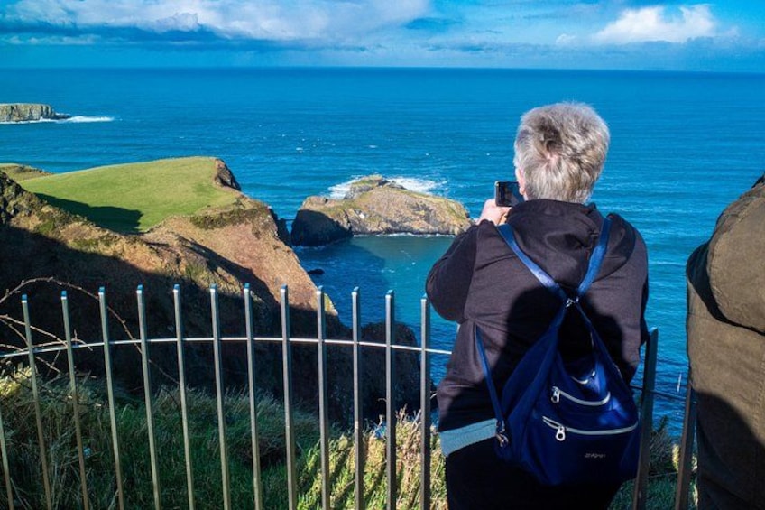 En person fotograferer havet fra en græsklædt klippe, med et metalhegn foran. 2