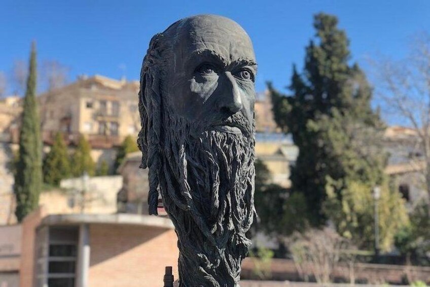 Una estatua con barba larga en un entorno exterior, con edificios y árboles de fondo. 3