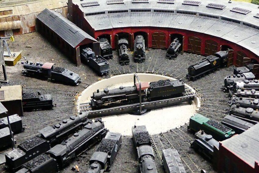 Un réseau de trains miniatures comprenant plusieurs voies et plusieurs trains, une plateforme circulaire centrale et un bâtiment rouge doté de nombreuses portes. 4