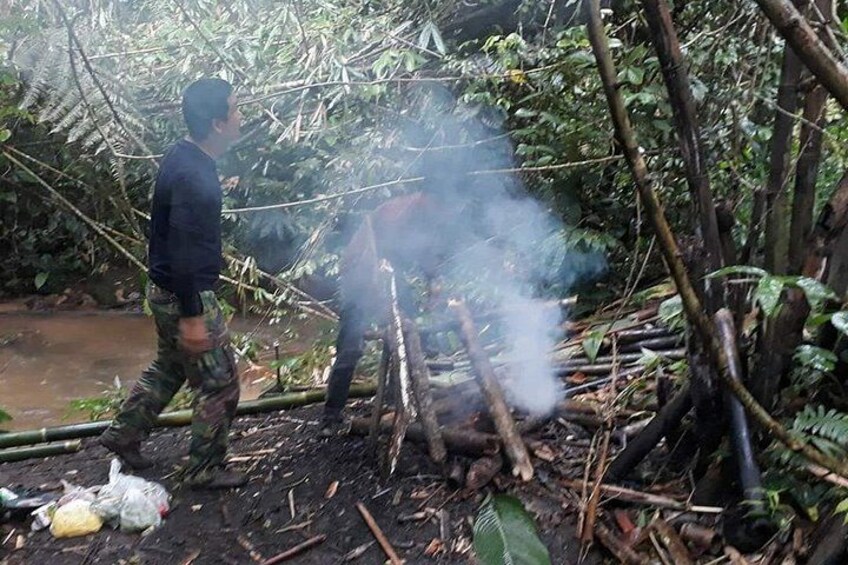 A person in camouflage clothing walking through a forested area with smoke rising from a fire. 1
