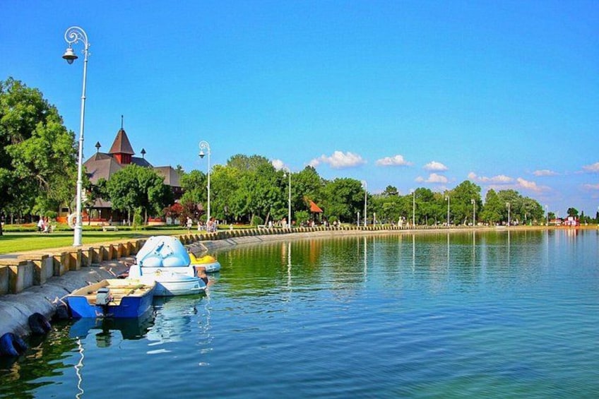 Ein ruhiger See mit vertäuten Booten links, ein Park mit Bäumen und ein Gebäude mit einem spitz zulaufenden Dach, und ein klarer, blauer Himmel. 3