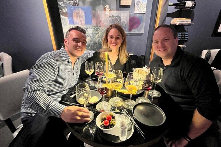 Three people seated at a table with wine glasses, plates, and a bowl of food. 1