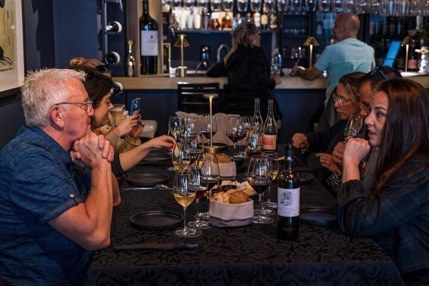 A group of people sitting at a table with wine glasses and bottles, in a bar setting with shelves of bottles in the background. 2
