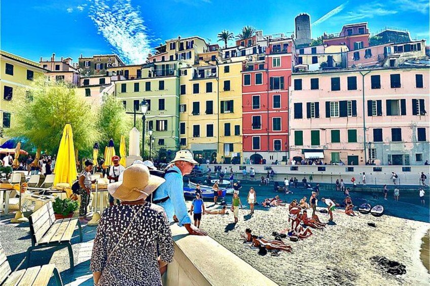 A crowded beach with people sunbathing and swimming, colorful buildings in the background, and a clear blue sky. 3