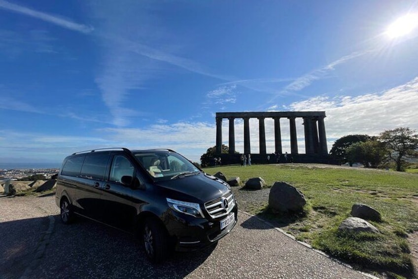 A black minivan parked on a paved path with a large classical building in the background. 1