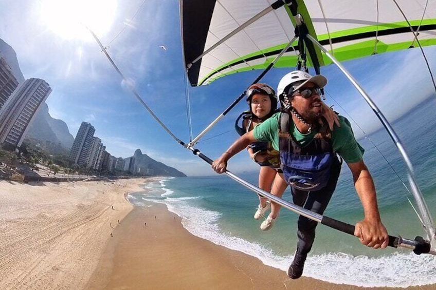 A person and a child on a hang glider over a beach with buildings in the background. 1