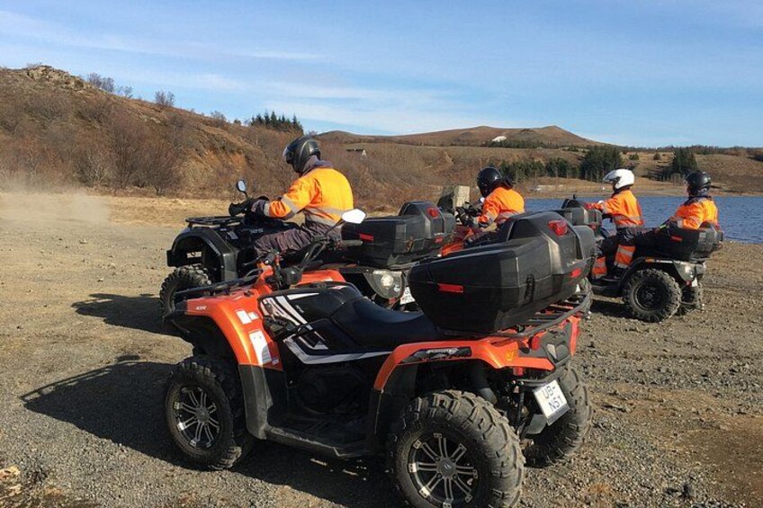 Four people on ATVs in a natural setting with a body of water and hills in the background. 5