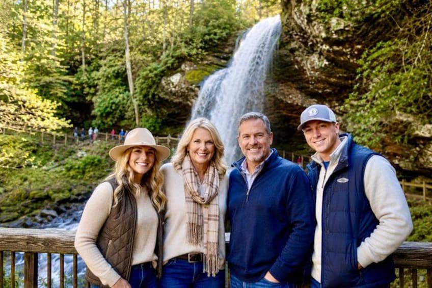 A group of people standing in front of a waterfall in a forested area. 1