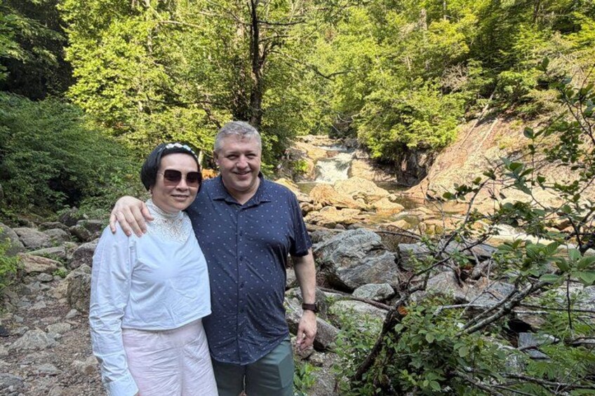 Two people standing in a forested area with a stream in the background. 3