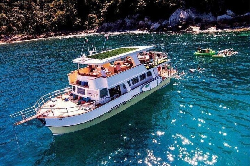 A white boat with a deck and lounge chairs on a turquoise sea, surrounded by rocky coastline and other boats. 1