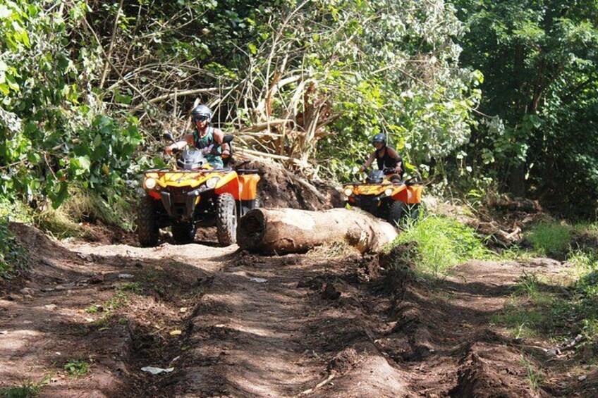 Two people on ATVs navigating a muddy off-road trail in a forested area. 3