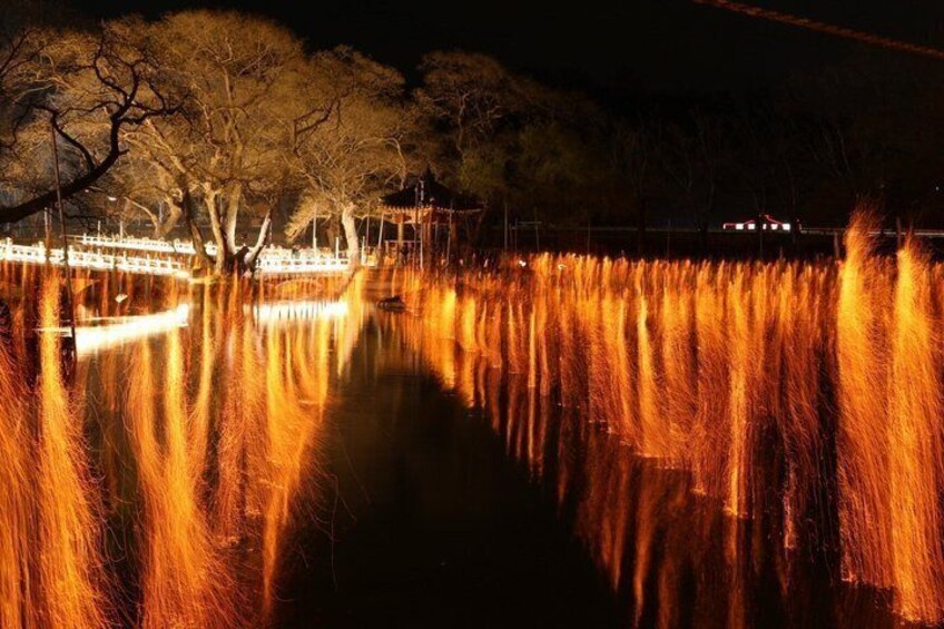 A nighttime scene with a body of water reflecting lights, trees, and a distant bridge. 2