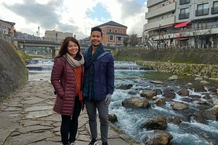 Una coppia in piedi vicino a un fiume che scorre, con un sentiero di pietra e edifici sullo sfondo. 4