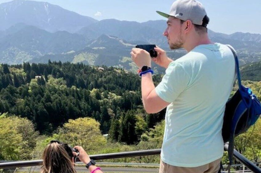 A person taking a photo of a mountainous landscape with dense forests and a clear sky. 5