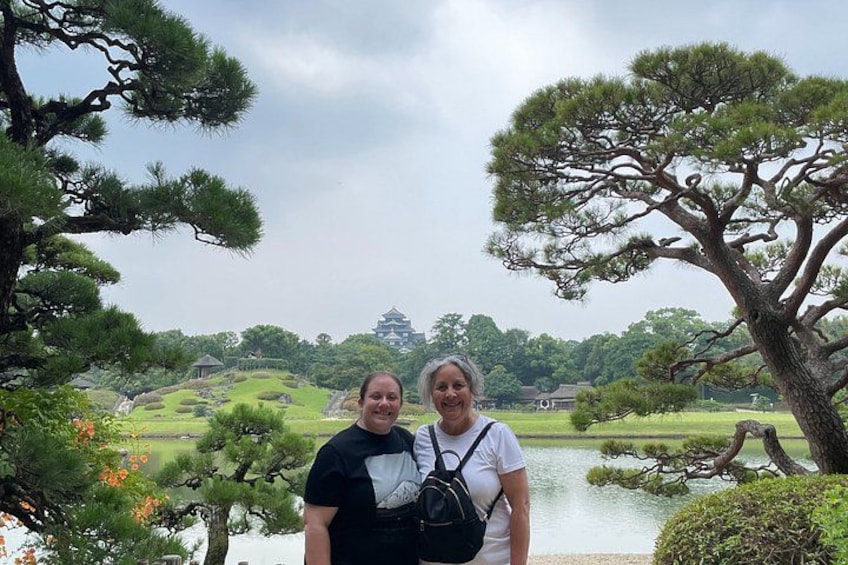 Two people standing in a park with a large tree, a pond, and a distant castle-like structure. 2