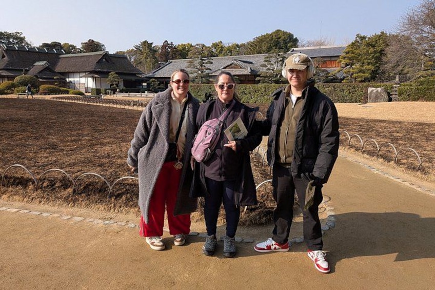 Three people standing on a dirt path with a traditional building and a fenced area in the background. 5