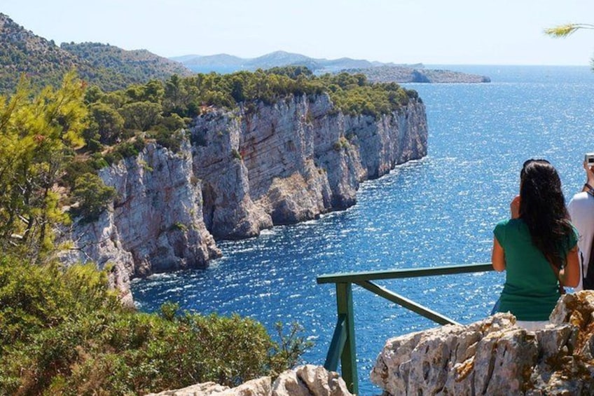 A coastal cliff with a railing, a person taking a photo, and a clear blue sea. 2