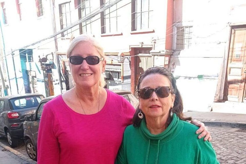 Two people standing on a cobblestone street with buildings and a parked car in the background. 3