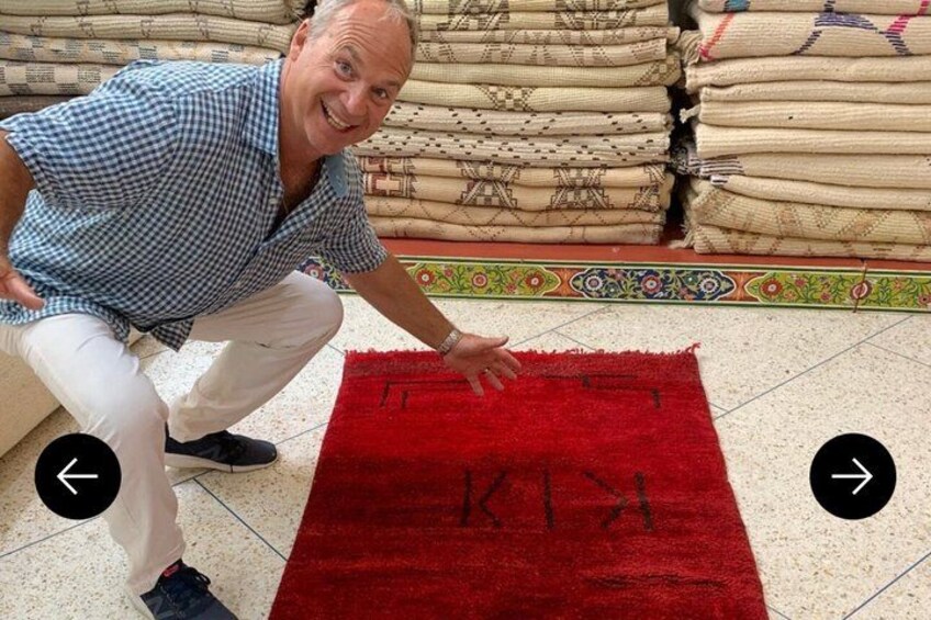 A person crouching next to a red rug with the word 'KIKI' on it, surrounded by stacks of beige rugs. 3