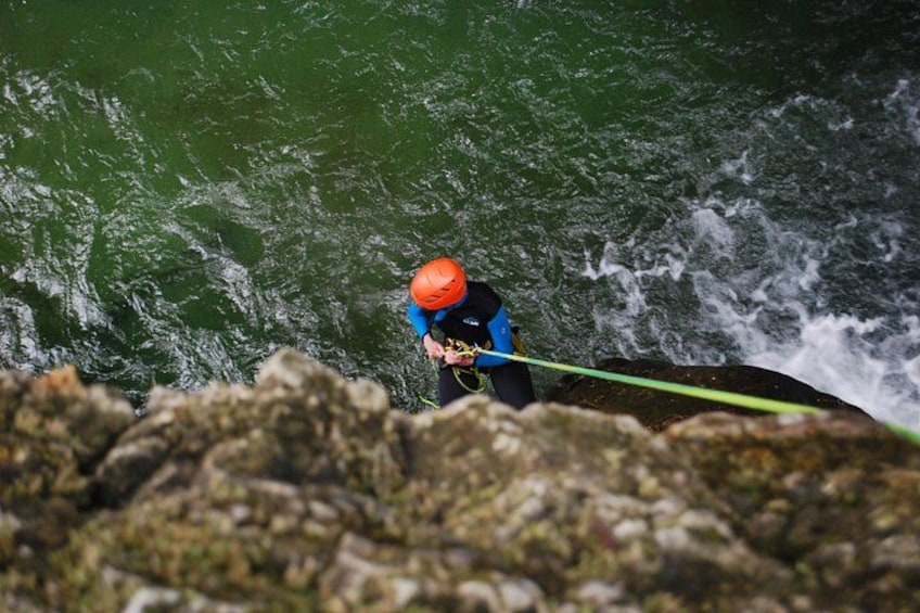 Una persona con traje de neopreno azul y casco naranja, sosteniendo una cuerda verde, sobre una superficie rocosa cerca de un cuerpo de agua. 3