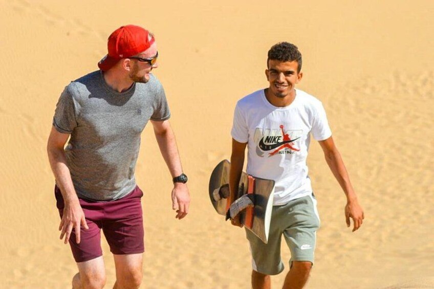 Dos personas caminando por un desierto con dunas de arena al fondo. 2