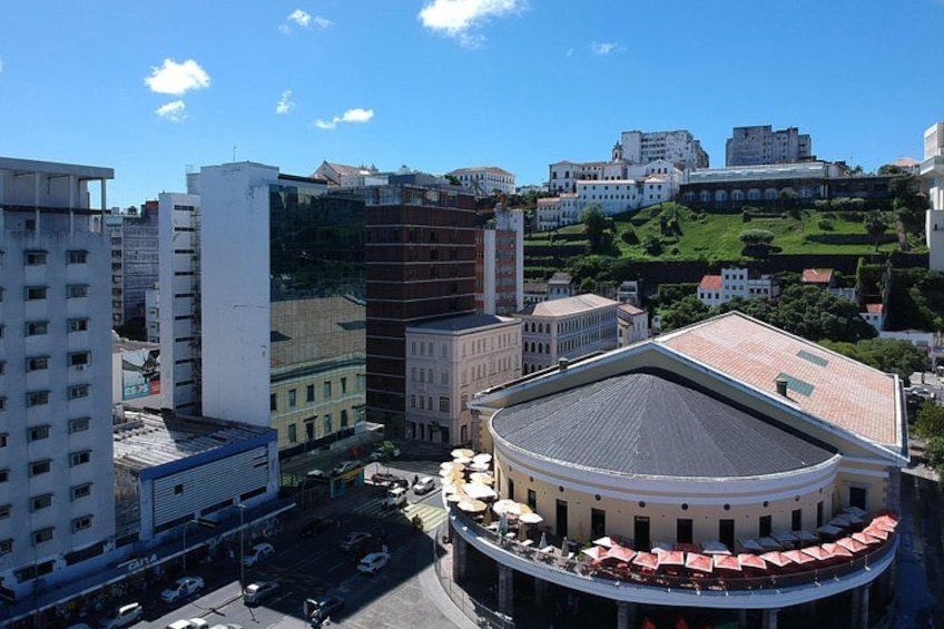 A cityscape with a prominent round building with a red roof, surrounded by modern high-rise buildings and a hill with greenery in the background. 5