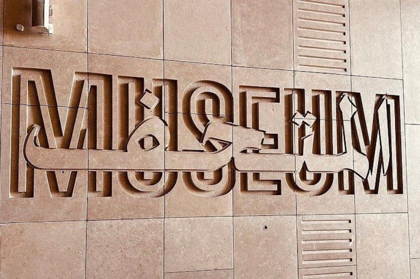 A large, three-dimensional sign with the word 'MUSEUM' in Arabic script, mounted on a tiled wall. 3