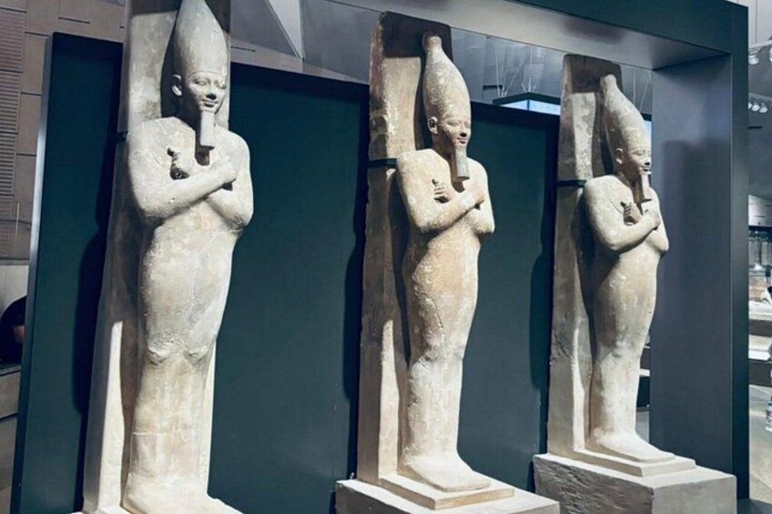 Tre antika stenstatyer med spetsiga hattar och korsade armar, utställda i en modern museimiljö. 2