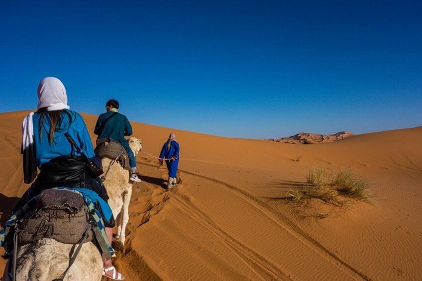 Drie mensen rijden op kameel door een woestijn met zandduinen en een heldere lucht. 2
