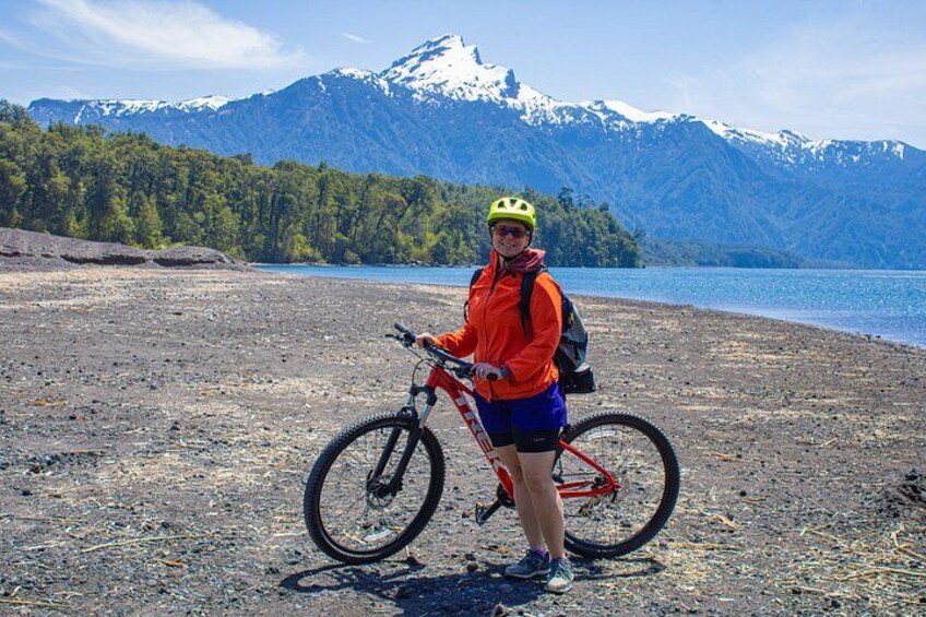 A person standing next to a mountain bike on a rocky beach with a lake and snow-capped mountains in the background. 5