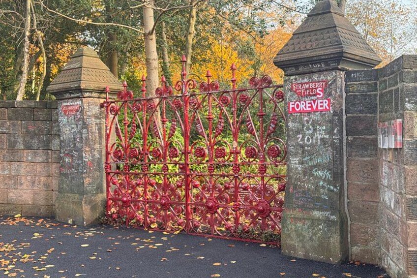 A red iron gate with a sign that reads 'Strawberry Fields Forever' and a brick wall with graffiti. 1
