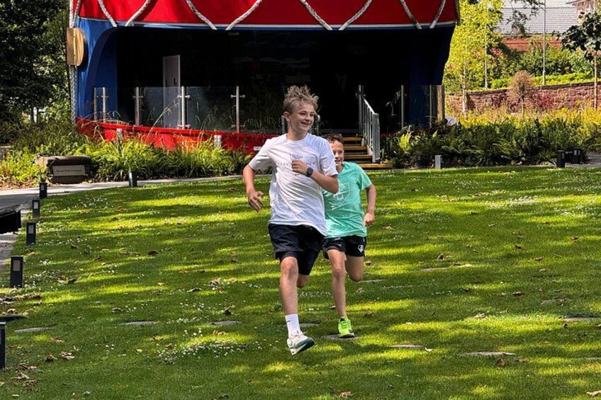 Two people jogging on a grassy field with a stage and trees in the background. 3